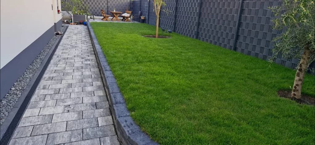 Gepflasterter Weg neben einer Rasenfläche und einem grauen Zaun